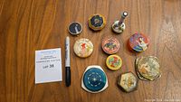 Top view of 10 vintage make-up compact cases with various vintage painted and enameled motifs along with a slim metal antique perfume atomizer standing upright on a table, next to a Sharpie and auction lot card.