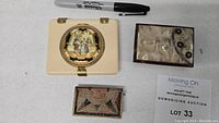 Three antique powder compact cases displayed on a white background with a black marker for scale and auction lot card.