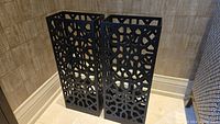 Pair of tall black stained wooden lattice candle lanterns with geometric pattern shown side by side against wall.