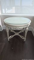Front view of round Louis XVI style table with carved apron and glass top against window