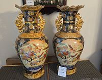 Pair of tall porcelain vases with detailed hand-painted geisha, birds and floral design with gold accents, placed on a table.