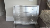 Front view of the silver painted chest of drawers showing three drawers with glass knob handles and curved front design.
