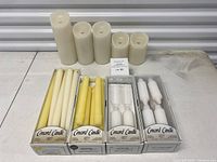 Five ivory battery-operated pillar candles of varying heights with some wear, behind four unopened boxed sets of Concord Candle taper and decorative candles in white and yellow.