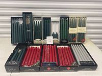 Multiple boxed sets and bundles of taper candles in green, red, and grey colors from brands Twilight and Crouse Candles, displayed frontally to show packaging and candle colors.