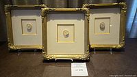 Three square gold-toned ornate frames with cream mats and central plaster cameos arranged side by side on stands.