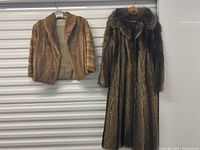 Two fur coats hanging on a wall, one short light brown coat and one long darker brown coat with fur collar.