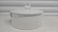 Front view of vintage white porcelain lidded casserole dish with basket-weave and vine embossed pattern, two side handles.