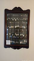 Full front view of the wooden spoon display case mounted on a wall, showing all souvenir spoons inside under glass.