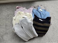 Seven women's winter sweaters in various colors and styles, folded and stacked on a light carpet.