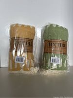 Two packaged Teema Turkish cotton towels or blankets, one mustard yellow and one olive green, both wrapped in plastic showing brand label.