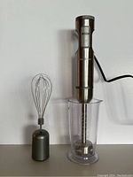 Hand blender in stainless steel with attached blending shaft in clear measuring cup and wire whisk attachment on side.