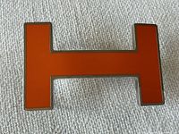 Front view of orange enamel H-shaped Hermès belt buckle on white background