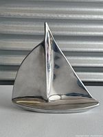 Front view of a polished pewter sailboat sculpture with clean modern lines and a large sail, placed on a gray surface with a corrugated metallic background.