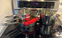 Pot and pan set neatly arranged on electric stove, showing three pieces with glass lids and one frying pan with red silicone handle cover