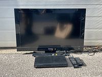 Sony 32 inch Bravia TV with DVD player, two remotes, and cords arranged outdoors on pavement.