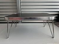 Side profile of a vintage black TV dinner tray with chrome trim and hairpin legs on a flat surface.