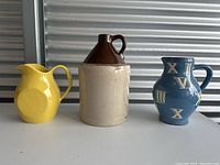 Front view of three pitchers and jugs; yellow ceramic pitcher, two-tone jug, and blue ceramic pitcher with Roman numerals