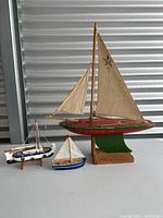 Front angle view of the three vintage wooden model boats showing largest Star sailing yacht with red and green hull and fabric sails, two smaller boats with cloth sails in blue and white paint.