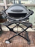 Full front view of Weber electric grill on foldable stand on balcony, showing closed hood, side shelves with brush, and power cord.