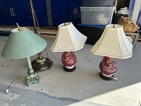 Three antique table lamps: two maroon ceramic lamps with cream lampshades and one green metal lamp with a green shade, displayed together on a concrete floor.