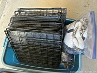 Multiple black metal wire grid panels stacked inside a blue plastic bin along with a sealed plastic bag containing fabric items, including a leopard print cat bed or blanket.