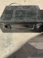 Front view of the black Denon amplifier showing volume and tuning knobs, digital display, input selector buttons, and headphone jack with surface dust and wear visible.