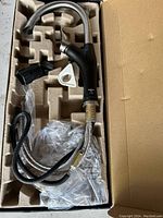 Blanco kitchen faucet with black body and stainless steel cobra head spout in packaging, showing attached braided water supply hoses and installation hardware.