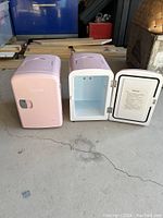Two pink mini fridges side-by-side on the floor, one closed and one open showing white interior with a small shelf.