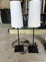 Pair of night table lamps with cords, white shades and black bases photographed on concrete floor.