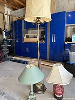 Photo of a tall antique floor lamp with two additional lamp shades placed near it. One cream colored shade with slight tear sits on top of lamp, and one green shade sits nearby. The lamp base is metal with a pullchain.