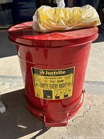 Red metal Justrite oily waste can with warning label and foot pedal, with yellow bio bags on top, placed on concrete floor.