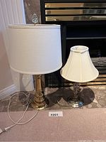 Two table lamps side by side on a tiled floor near a fireplace. One lamp gold with large round white shade, the other silver with smaller pleated white shade.