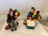 Both the male and female Royal Doulton Old Balloon Sellers figurines together, front view showing detailed colorful balloons and painted clothing.
