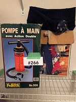 Photo showing boxed double action hand pump labeled 'Pompe à Main avec Action Double' by World Famous (No. 309) along with a folded dark-colored air mattress and a box with accessories.