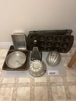 Photo of set showing aluminum bundt pans, muffin pan, baking pans, cooling racks, and grater on tiled floor.