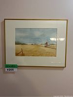 Framed watercolor painting of a Saskatchewan field featuring distant grain elevators, white mat, and gold frame, with visible reflection of ceiling lights.
