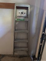 Close-up photo of a white metal ladder showing four steps and a top platform with holes, leaning against a wall in a garage.