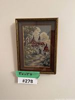 Framed needlepoint wall art showing a castle scene in muted tones with trees and clouds, in an ornate brown frame; image taken straight-on
