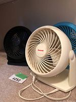 Three small desk fans arranged on a carpet, showing white Honeywell in front with black and blue fans behind.