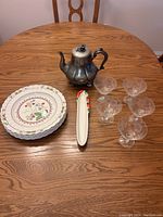 Overview of all items on dining table: 4 Spode plates stacked, 6 stemmed glass dessert bowls arranged, silver-toned Totley Sheffield tea pot, white ceramic leaf-shaped dish with flower and berry detail.