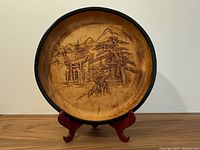 Front view of round wooden bowl with carved Japanese landscape, including deer, shrine gates, pine trees and mountains, black rim visible, placed on wooden stand.