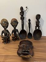 All seven African wood carvings including mask, male/female figurines, and three wooden spoons with figural handles arranged on a table.