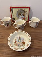Front view of three two-handled Bunnykins cups arranged in a row with a rimmed child's bowl and boxed gift set behind