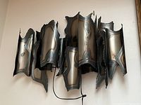 Front view of the vintage metal sculpture wall lamp mounted on wall, showing dark oxidized metal panels with wave-like edges and textured surface.