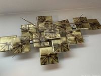 Front angle view of the metal sculpture showing the layered squares and rods in brass with a starburst pattern.