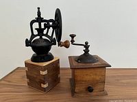 Pair of two vintage coffee/spice grinders shown side by side on wooden surface. The older wooden grinder with drawer on right, newer grinder with iron wheel on left.
