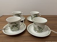 Four tea cups with matching saucers and spoons arranged on a wooden surface, showing green naive style garden and figure pattern.