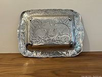 Polished aluminum serving tray with embossed rabbits and leaf design on wooden surface.