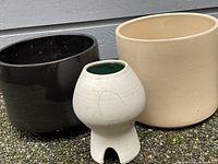 Three planters including two large vintage Gainey planters (one black, one beige) and a smaller unique handmade white planter with green interior, all placed outdoors on pebbled ground.