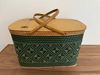 Full view of green woven picnic basket with wooden lid and two handles, showing detailed diamond pattern on side.